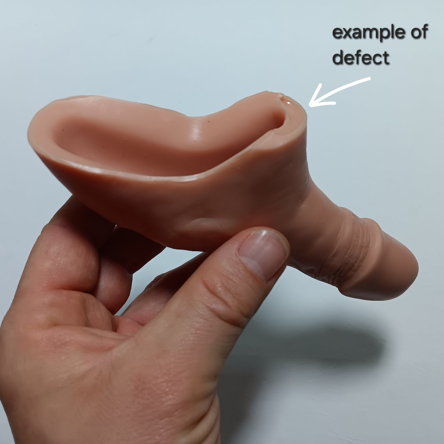 Hand holding a silicone product with an 'example of defect' label pointing to the rim where there is a defect and the top of the rim is cut short.