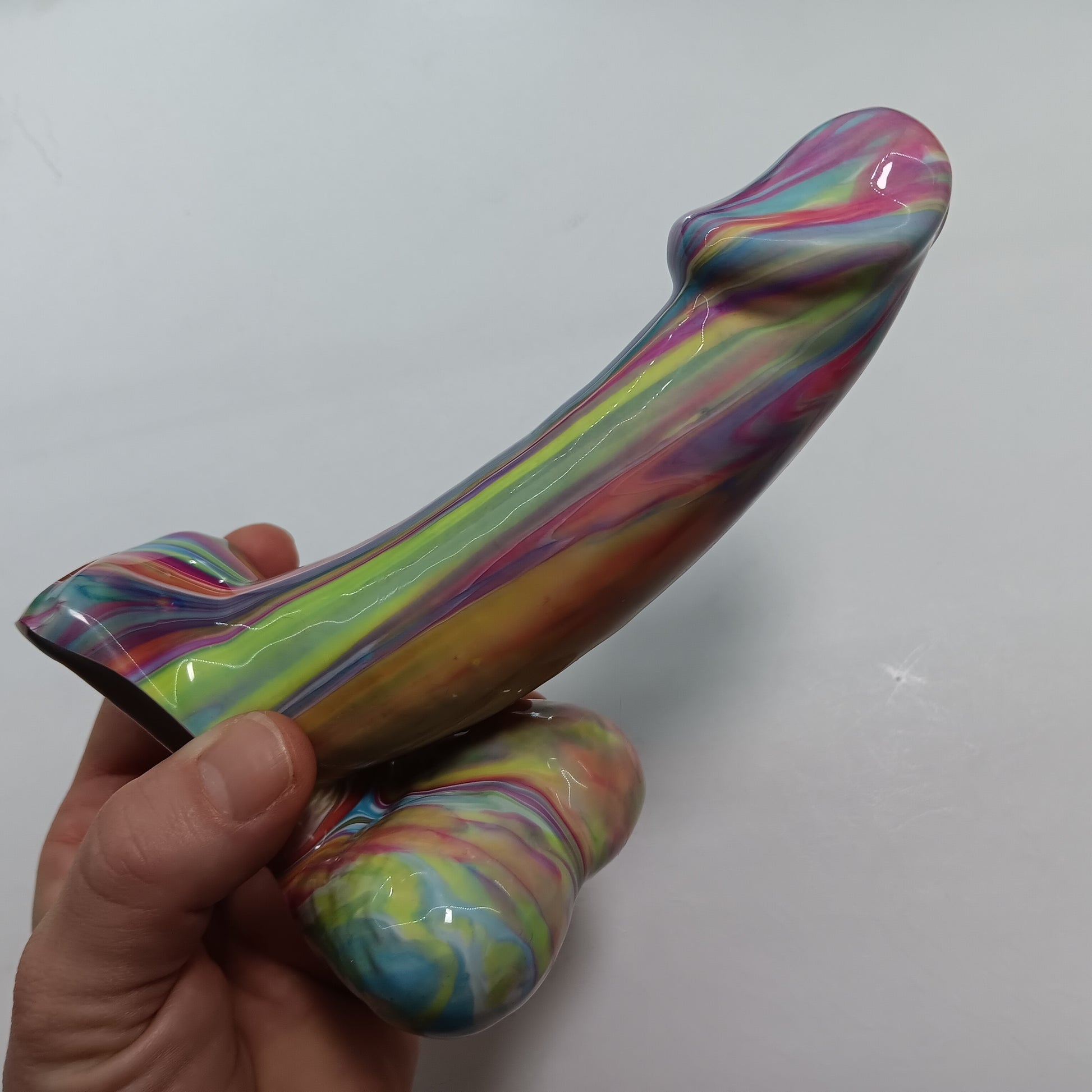 Hand holding the color drip solid toy- it is a shiny toy and the colors are pink, yellow, blue, purple, white, down the sides with a marble smooth appearance.  